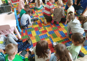 Zdjęcie przedstawia grupę dzieci siedzących na dywanie, które z wykorzystaniem tabletów starają się poruszać robota.